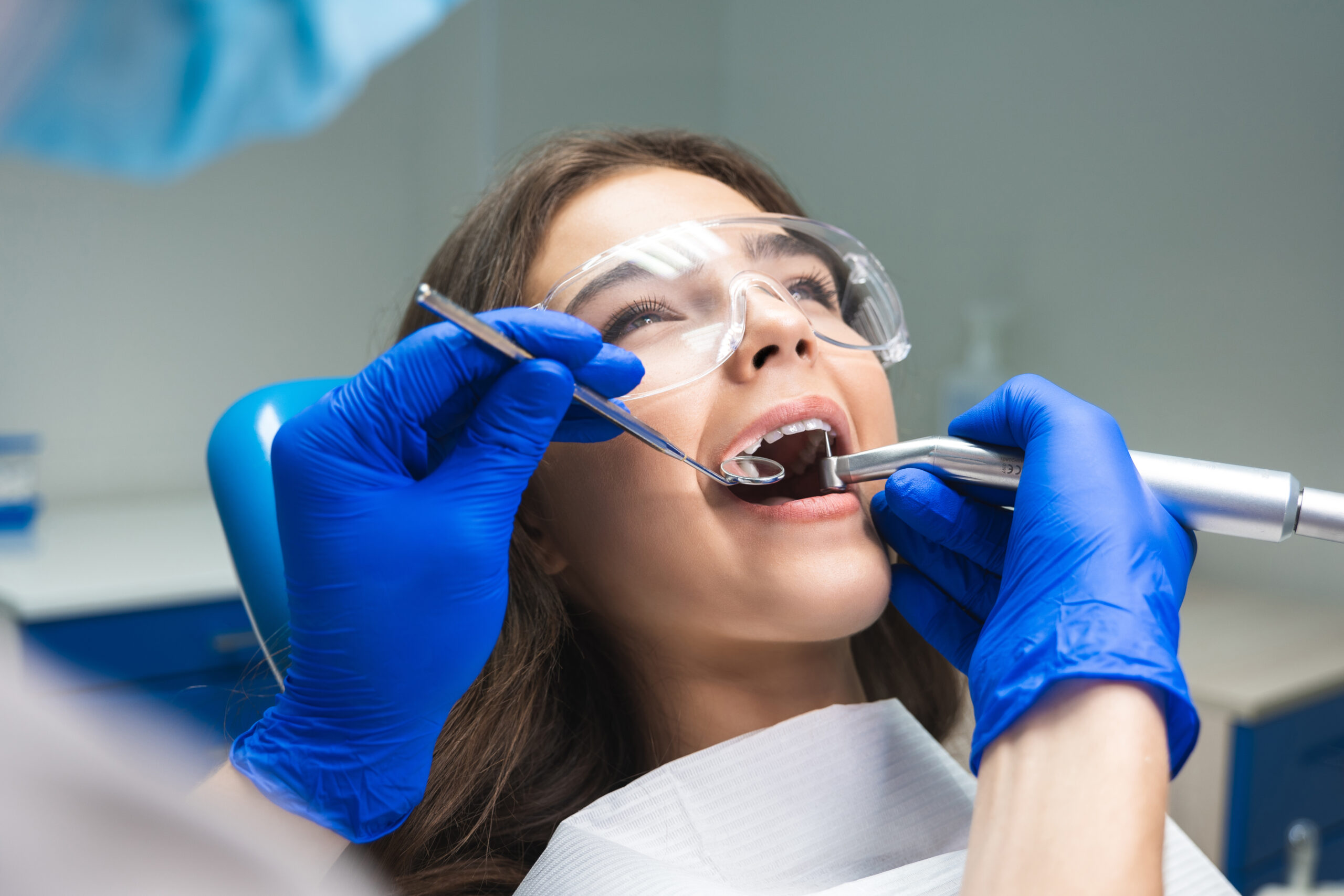 dentist in mask filling the patient's root canal while she is lying on dental chair wearing safety glasses under the medical lamp in dental office.